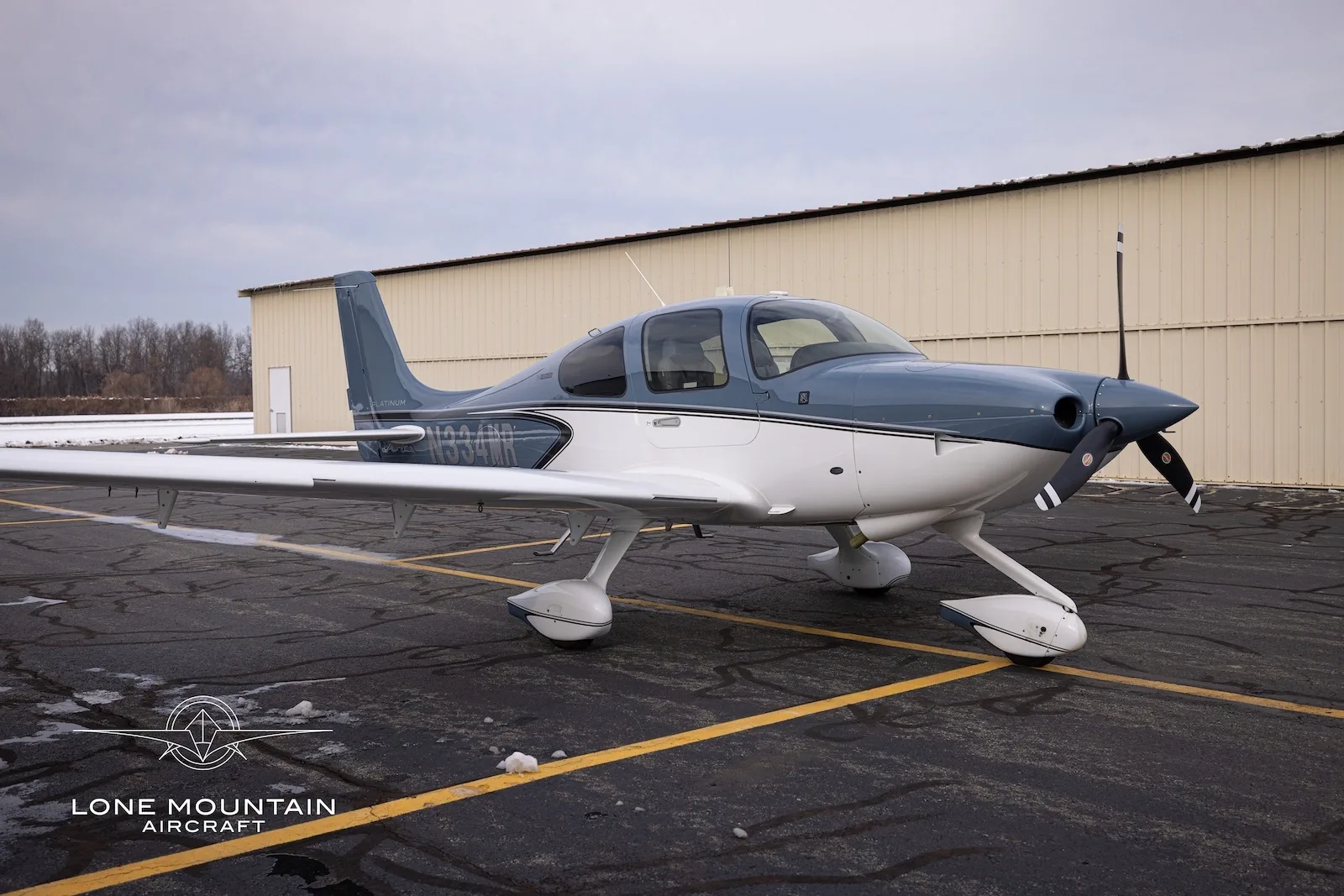 A Cirrus SR20 aircraft, N334MR, with a blue and white paint scheme, parked on a tarmac with some snow.