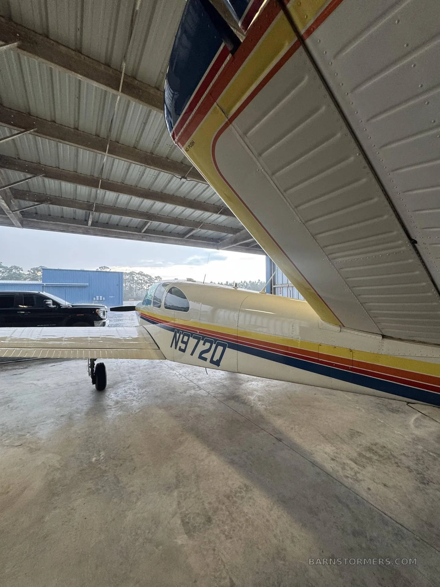 A close-up side view of a white aircraft with rainbow stripes and yellow wings, parked in a hangar with the door open to the outside.