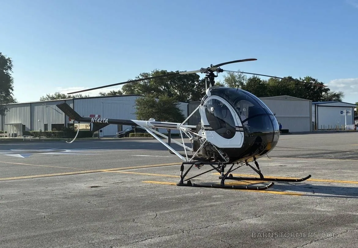 A black and white Schweizer 300CBi helicopter (N142TA) parked on an asphalt tarmac with hangars in the background.