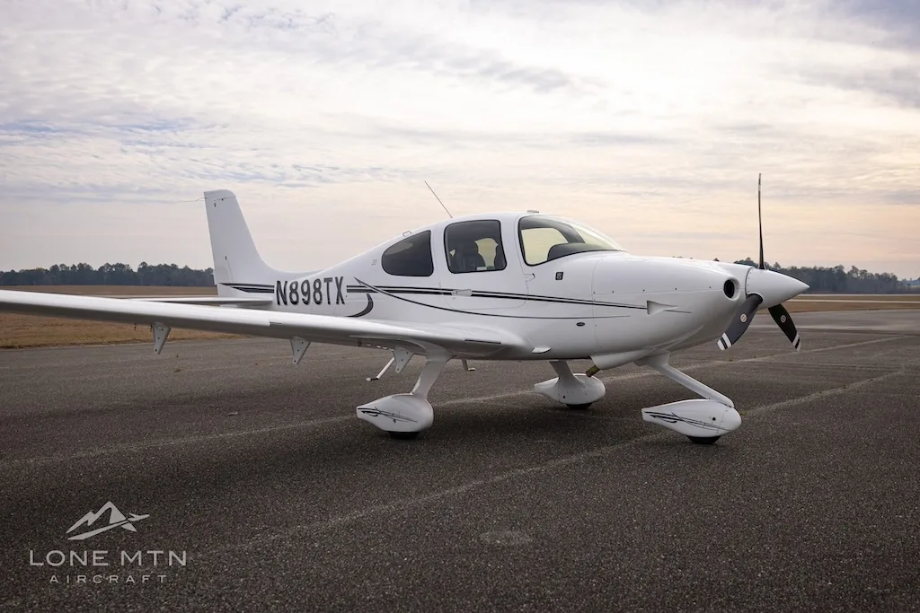 A white Cirrus SR20 aircraft with black and grey stripes, registration N898TX, parked on an asphalt runway under a cloudy sky.
