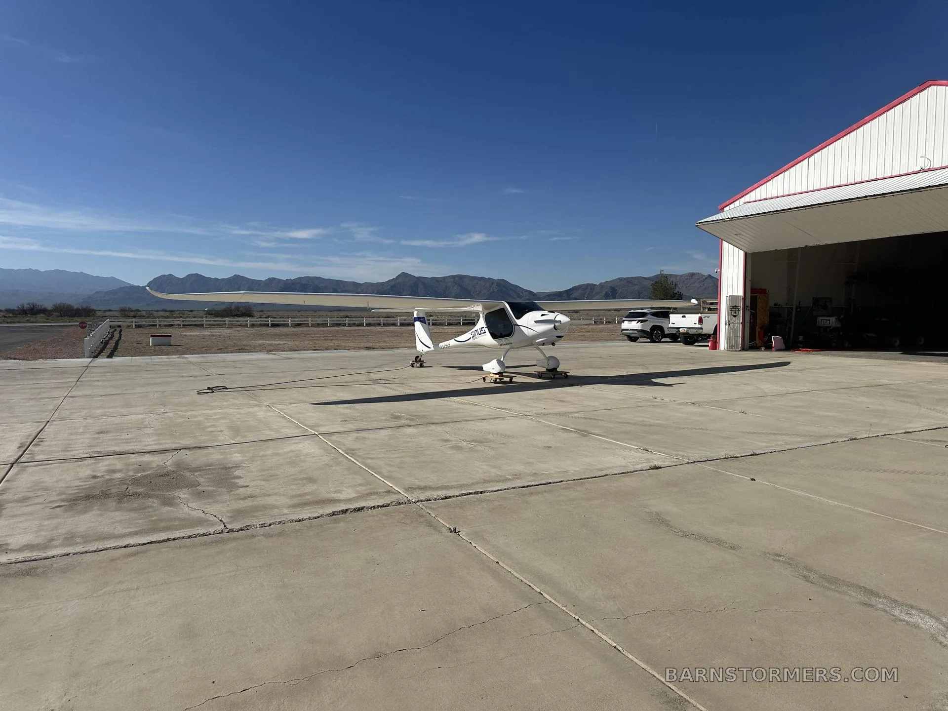 A white Pipistrel Sinus aircraft with blue accents is viewed from the front-right, parked on an apron with mountains in the background.