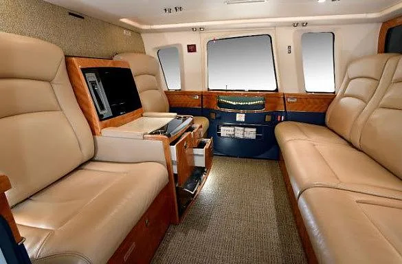 The spacious interior cabin of a helicopter with tan leather seating, wood accents, and large windows.