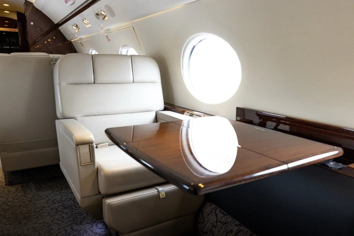 A cream leather passenger seat with a polished wooden table extended, next to a large oval window in an aircraft cabin.