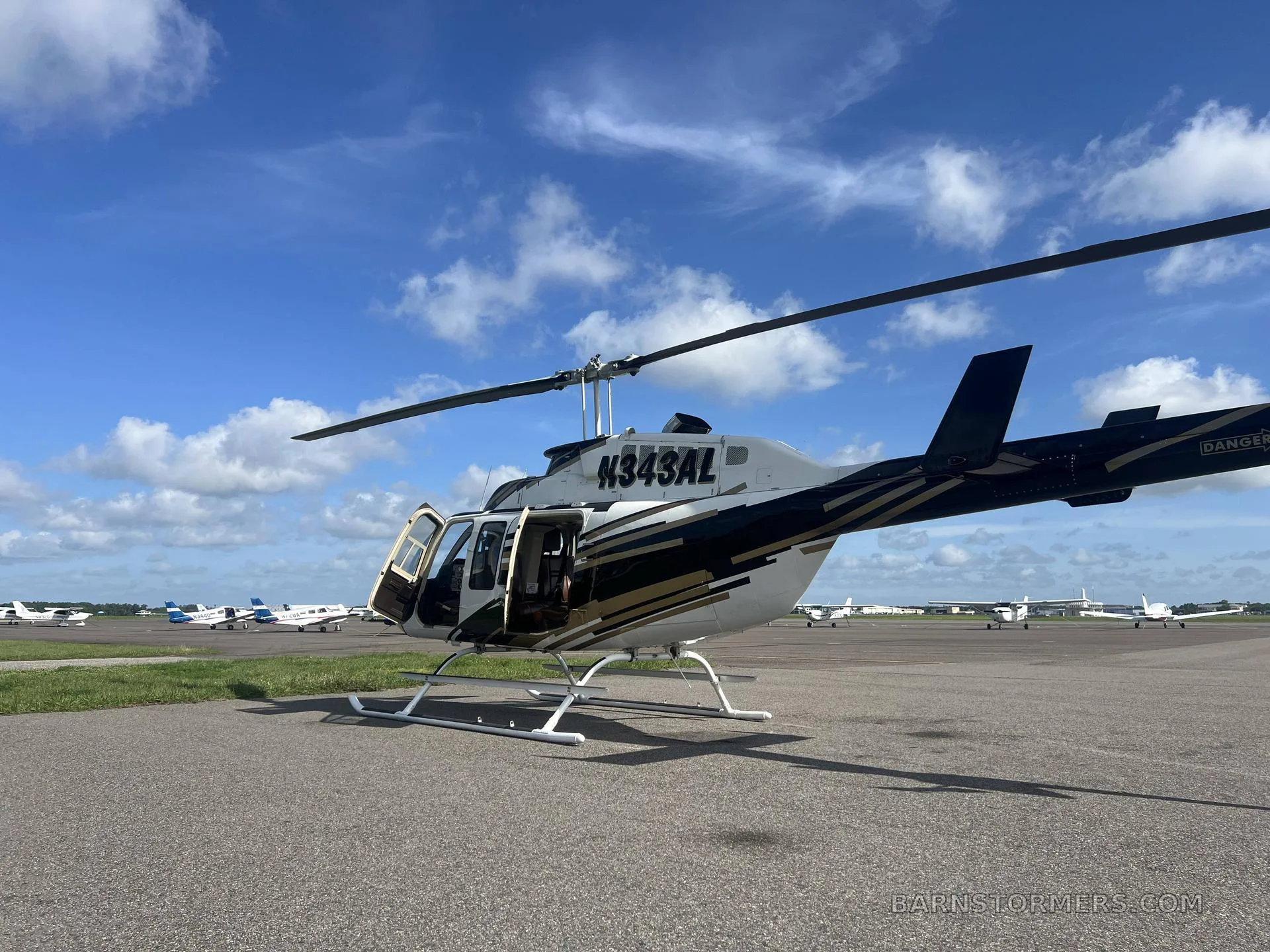 A white and gold Bell 206 helicopter (N343AL) with open doors is parked on an airport tarmac with other small aircraft in the distance.