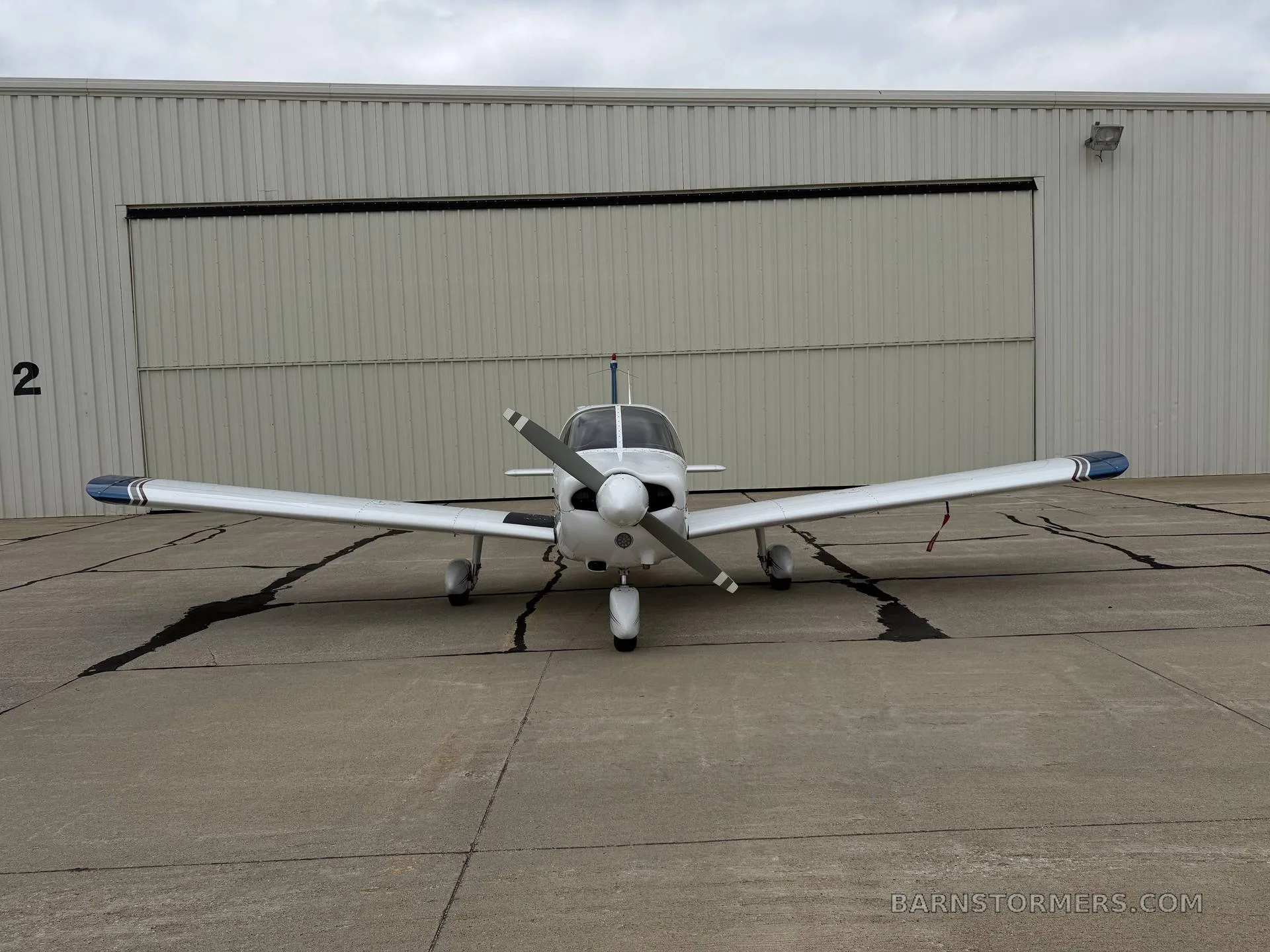 A white Piper Cherokee aircraft with blue accents, viewed from the front, parked on an asphalt apron in front of a hangar.