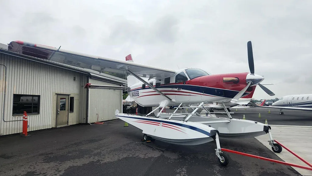 A red, white, and blue Quest Kodiak 100 on amphibious floats parked on an asphalt apron next to a hangar.