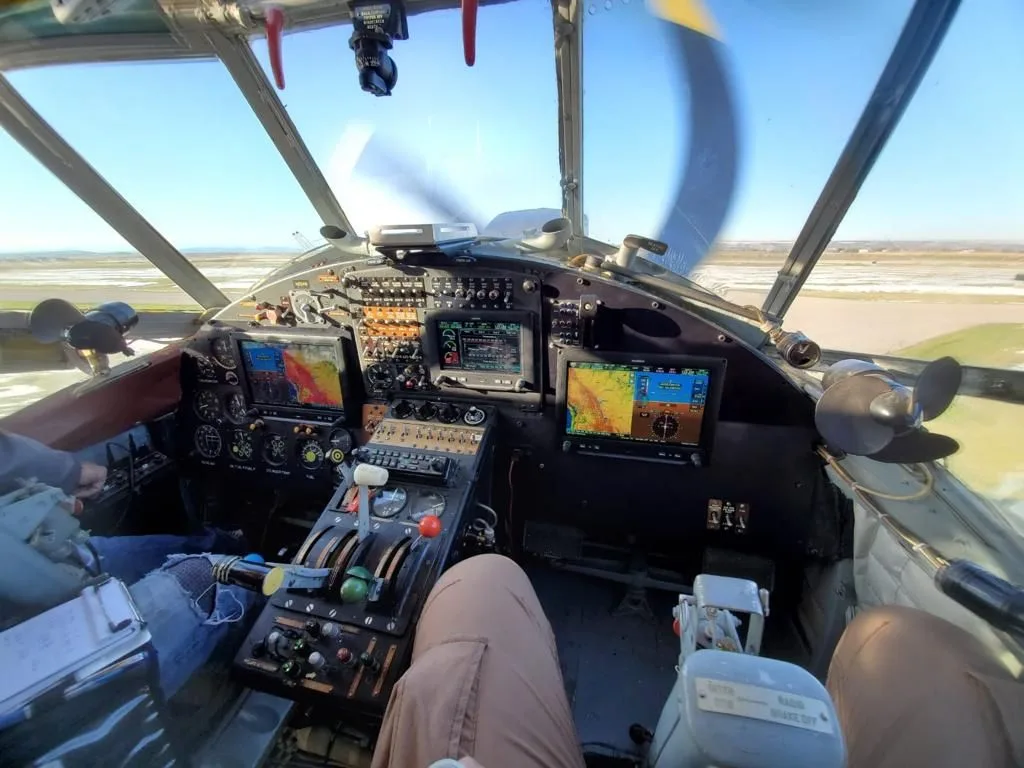 A wide view of the Antonov AN-2 cockpit from the pilot's perspective, showing multiple displays and controls with a spinning propeller outside.