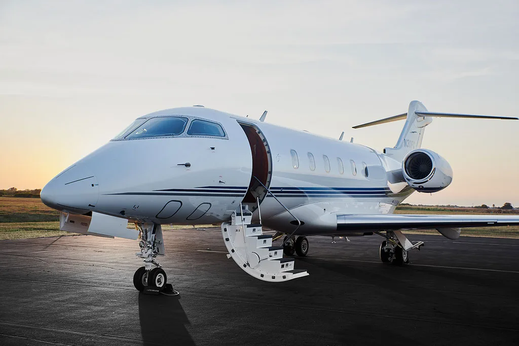 A white Bombardier Challenger 300 private jet with blue stripes and an open entry door on an airfield at sunset.