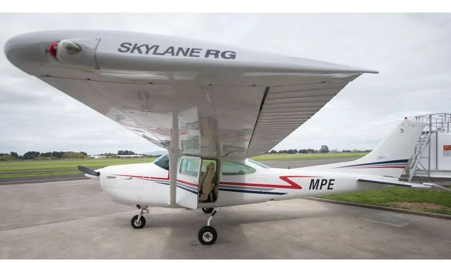 A white Cessna Skylane RG aircraft with red and blue stripes and 'MPE' on the tail, parked on a tarmac with its door open.