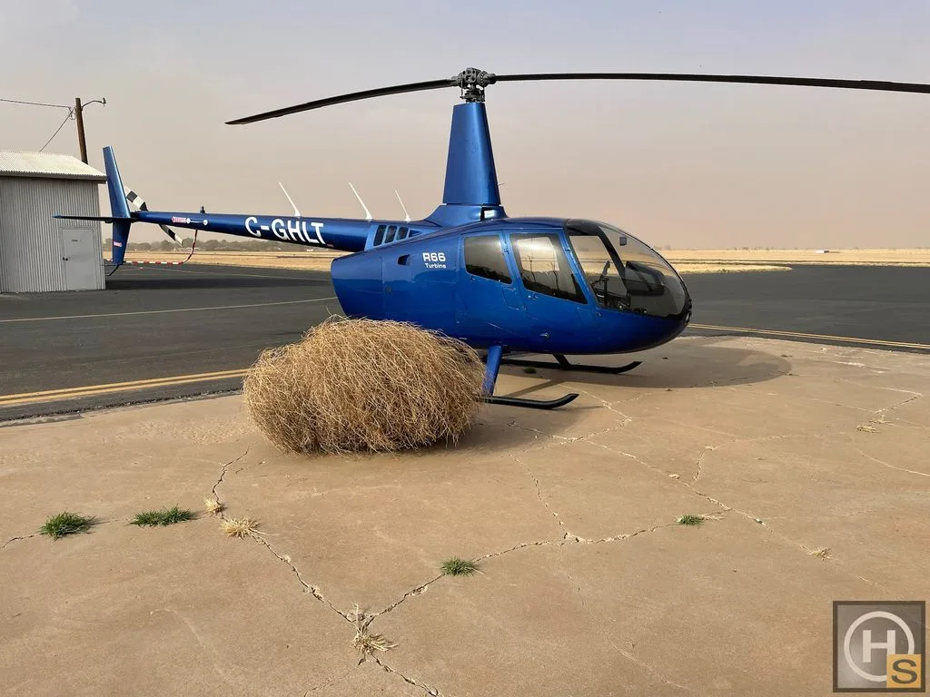 A blue Robinson R66 Turbine helicopter, registration C-GHLT, is parked on a concrete pad next to a tumbleweed.