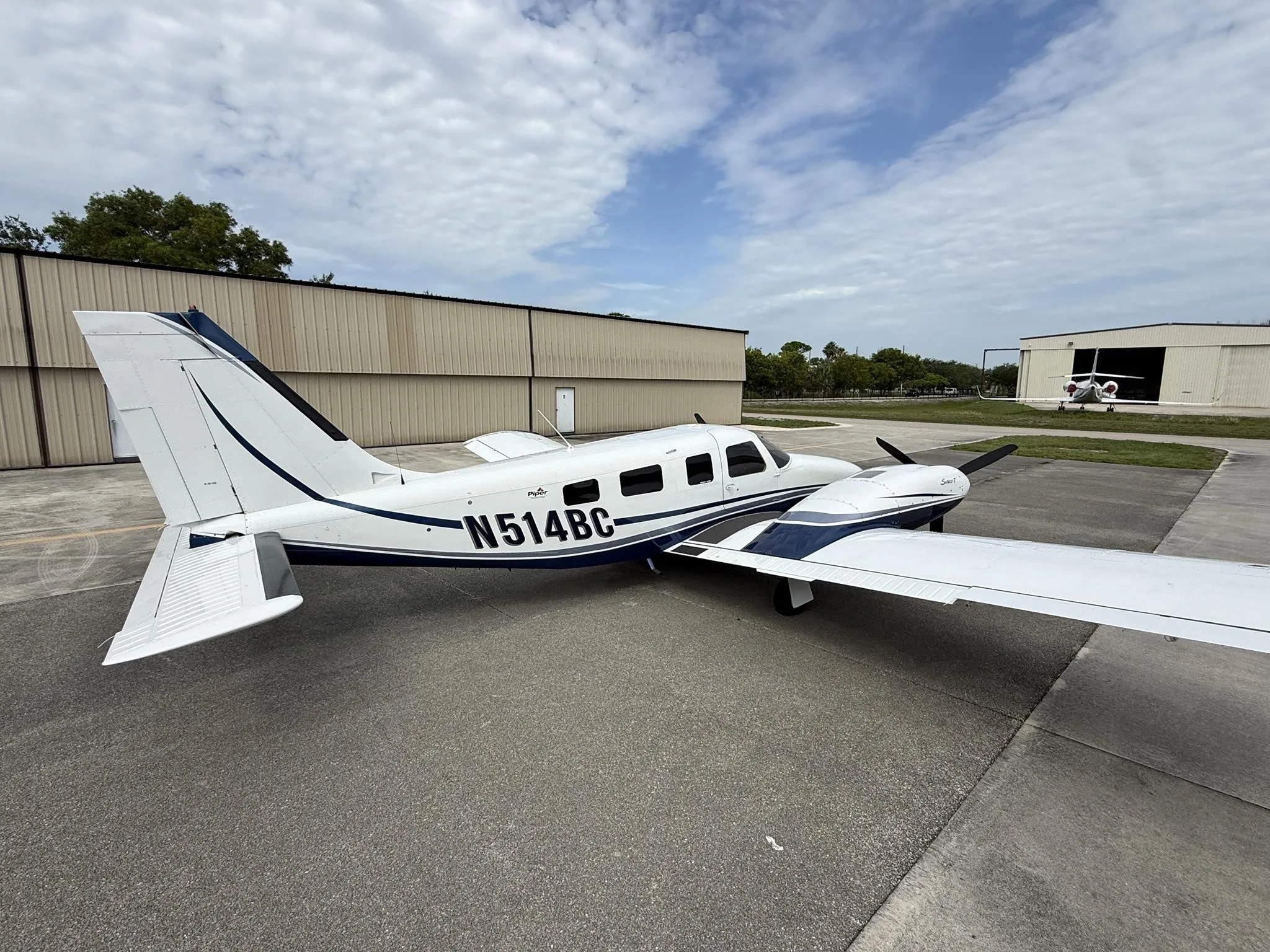 A white and blue Piper Seneca V aircraft, registration N514BC, parked on an airfield tarmac with hangars in the background.