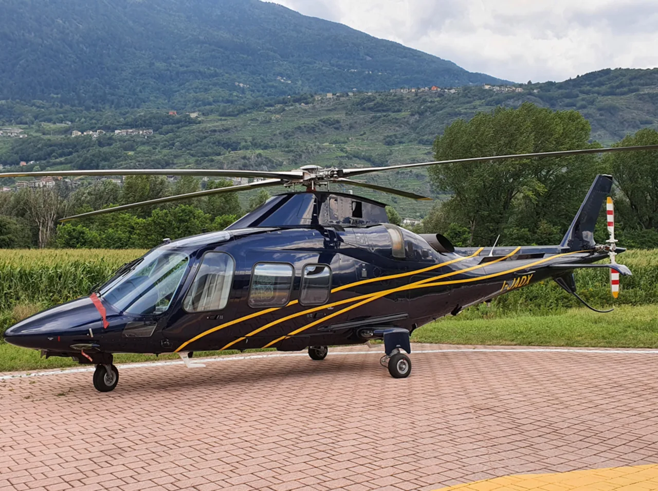 A dark blue AgustaWestland AW109 helicopter with yellow stripes is parked on a paved helipad with mountains in the background.