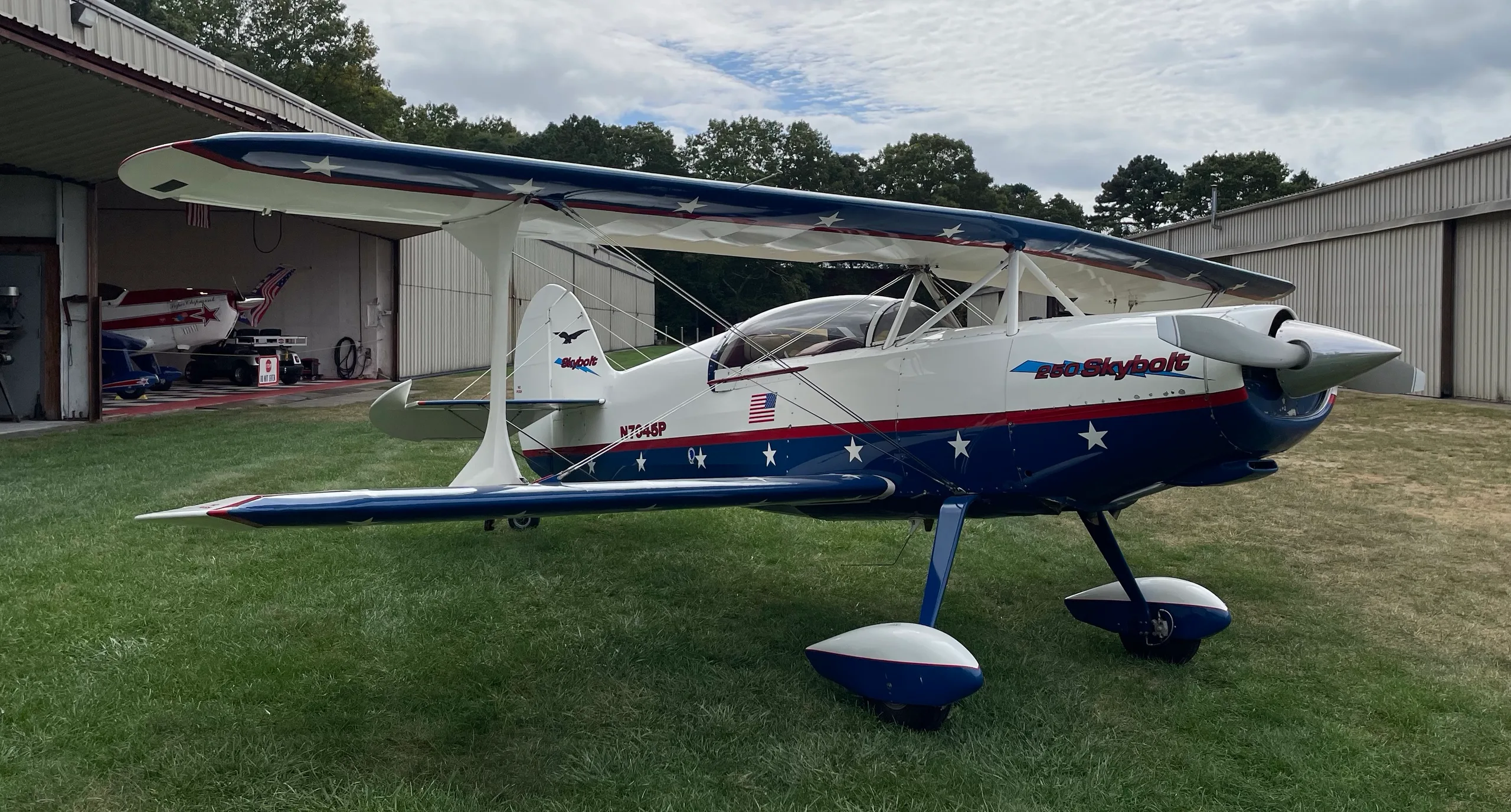 A white and blue Acro Sport II Skybolt biplane with red and white stars on the fuselage and wings, parked on grass next to a hangar.