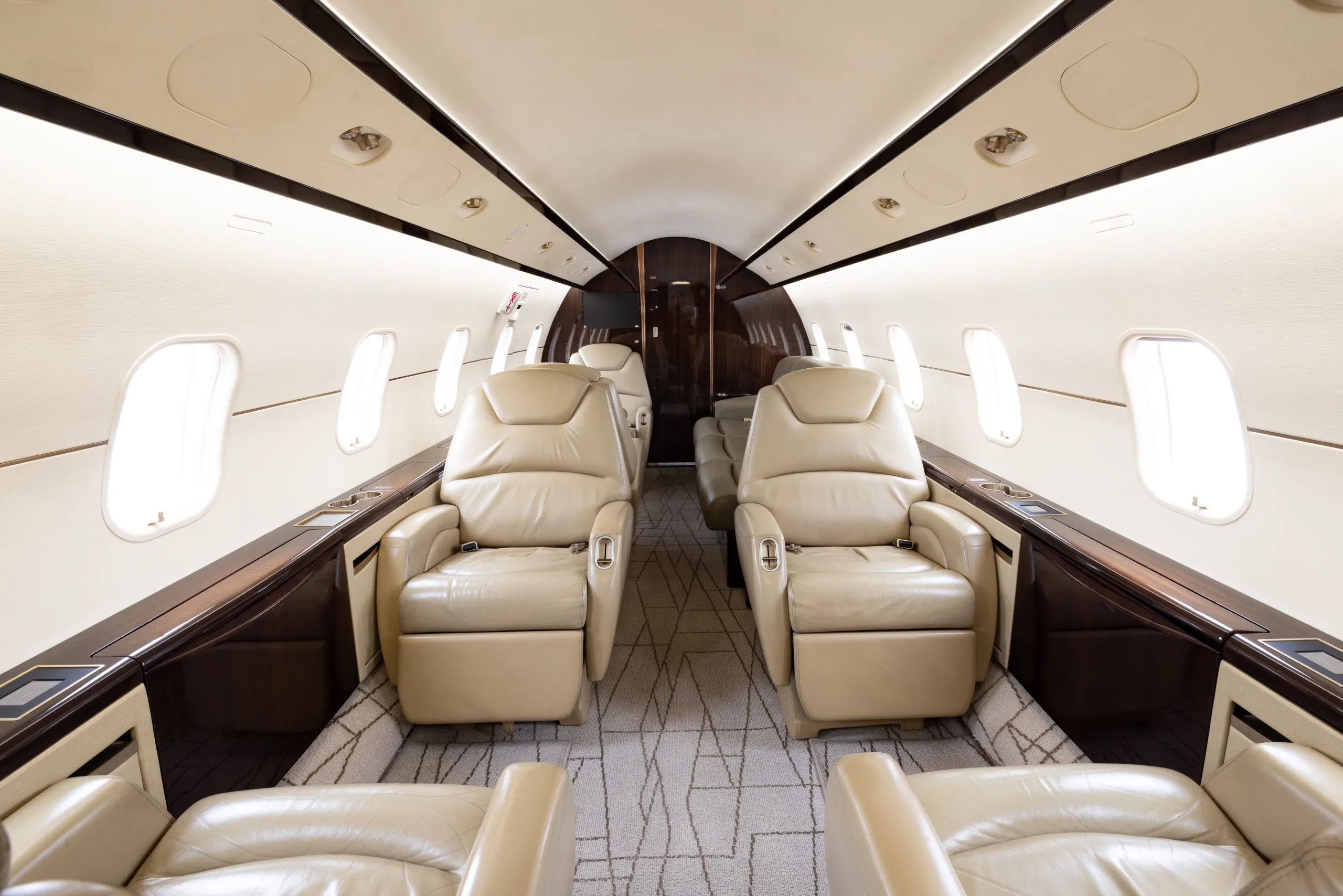 The luxurious interior of a business jet cabin with light beige leather seats and dark wood tables.