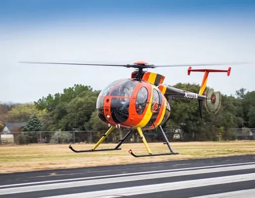 A Hughes 369HS helicopter with orange, yellow, and black stripes hovers above a runway.