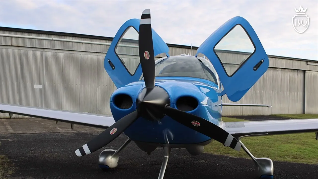A blue single-engine propeller aircraft with open gull-wing doors, parked in front of a hangar.