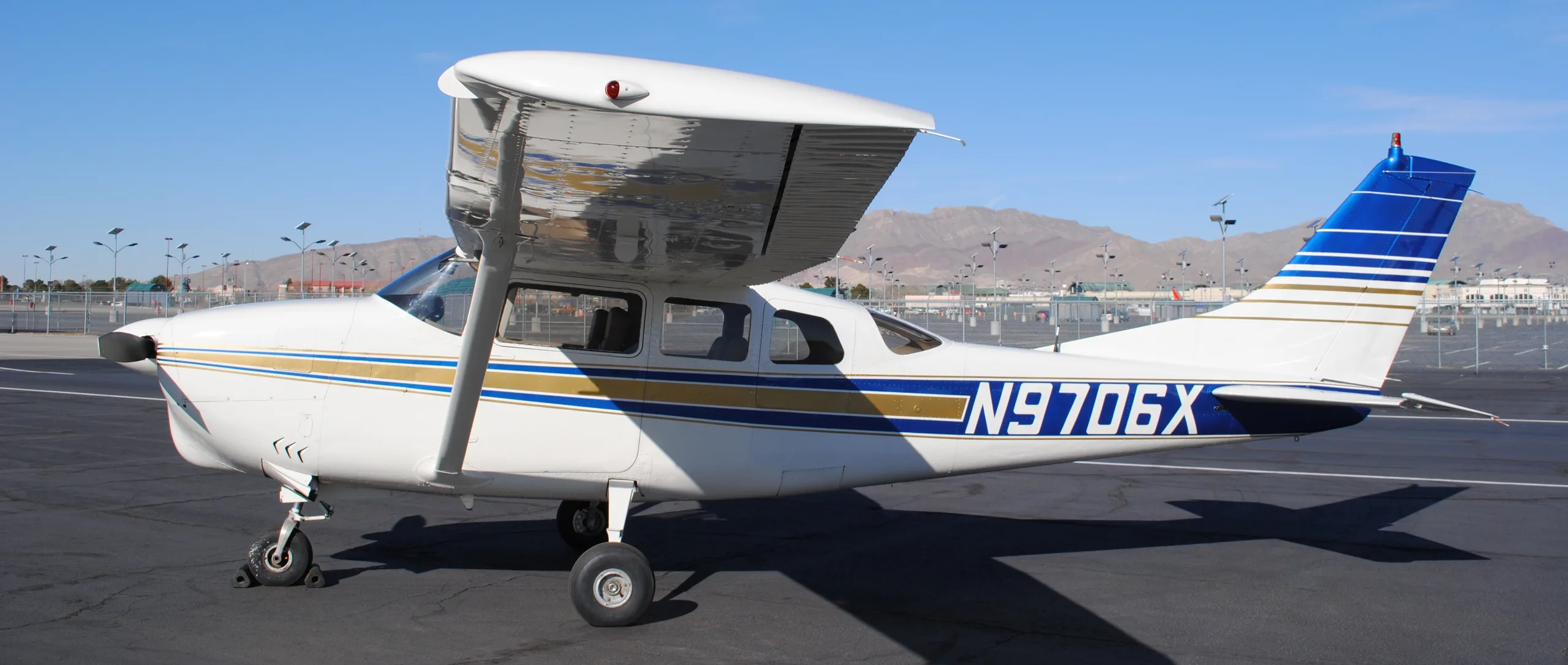 A white single-engine propeller aircraft with blue and gold stripes, tail number N9706X, parked on an asphalt tarmac with mountains in the background.