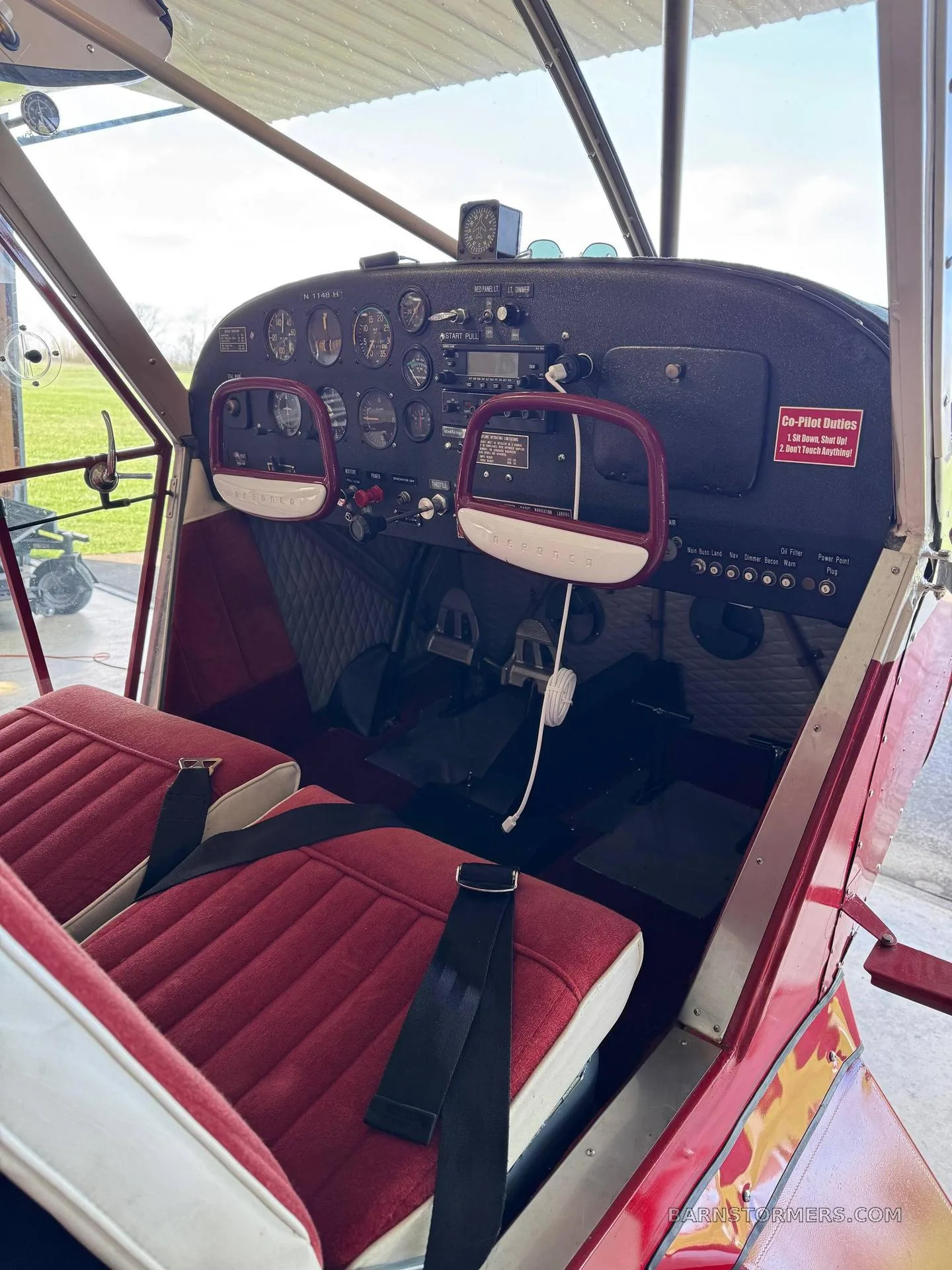 The cockpit of a red and white vintage aircraft, showing the instrument panel, yokes, and red upholstered seats.