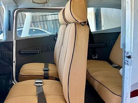 The interior cabin of an aircraft with tan seats and black piping, showing the rear passenger area.