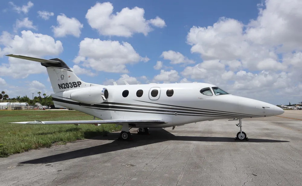 A white Embraer Phenom 100 business jet with black and gold stripes on the fuselage and tail, parked on an asphalt ramp under a partly cloudy sky.