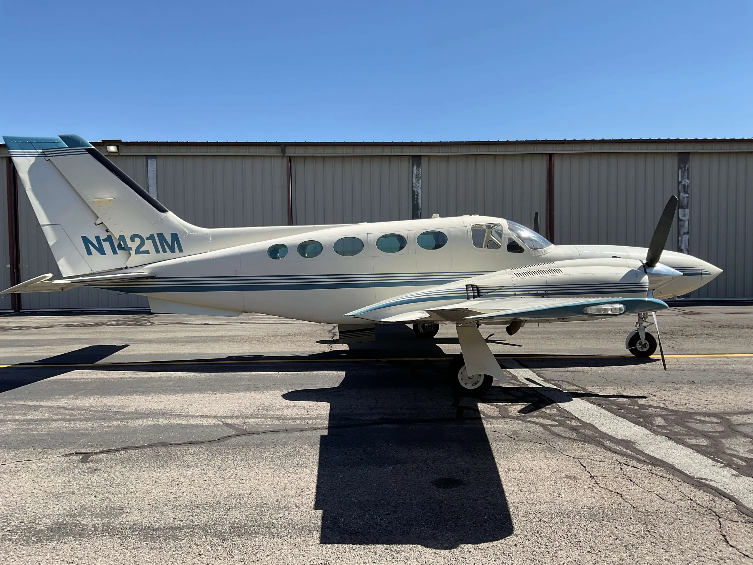 A white twin-engine propeller aircraft with blue stripes, tail number N1421M, parked on an asphalt ramp in front of a hangar.