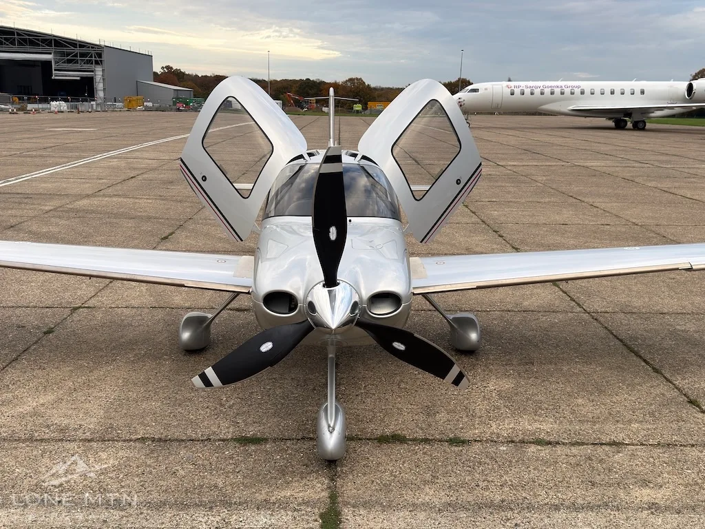 A silver Cirrus SR22T aircraft with open doors viewed from the front on an airfield tarmac.