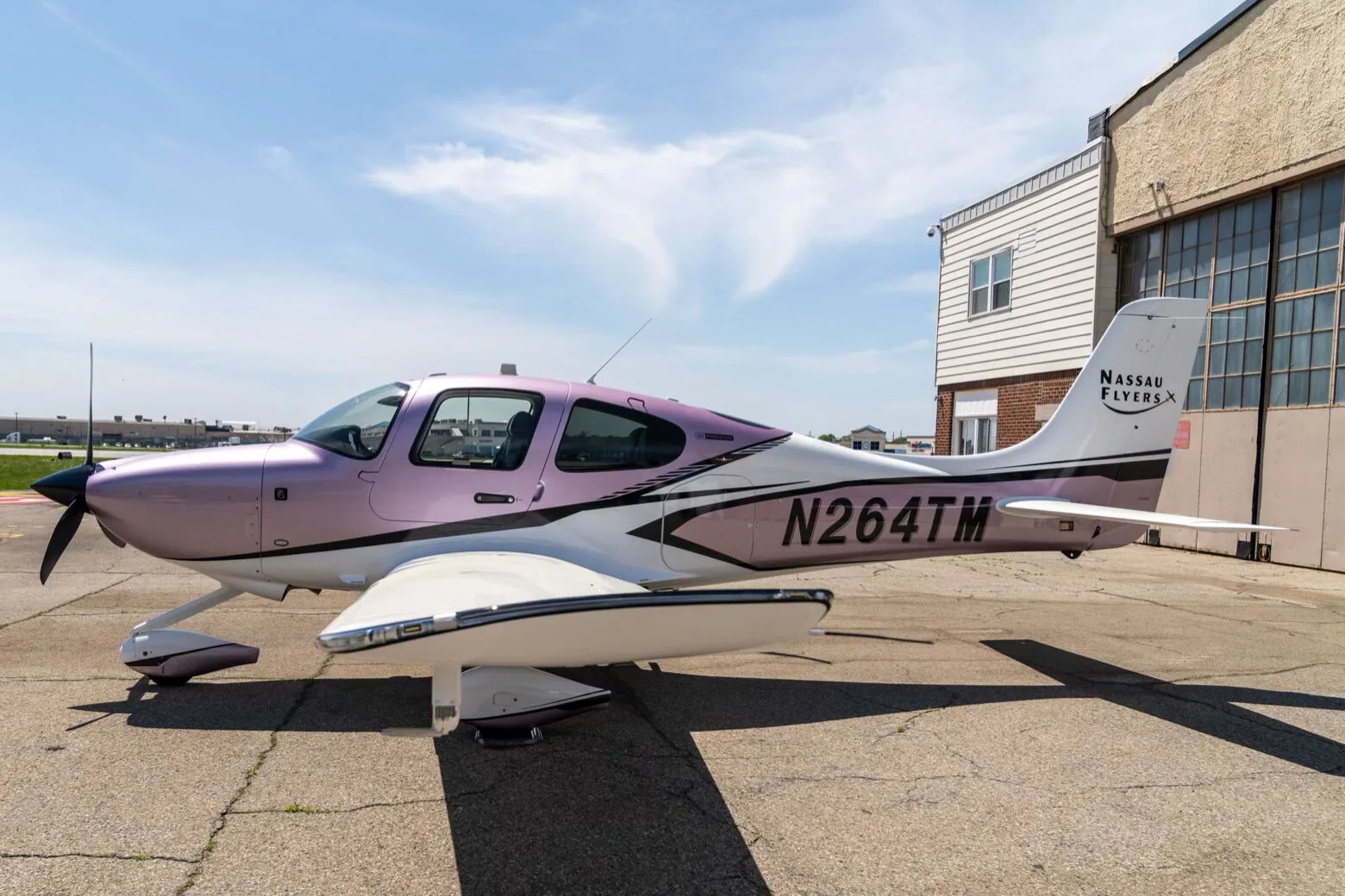 A Cirrus SR22 aircraft with a metallic lavender and white paint scheme, registration N264TM, parked on an airfield next to a building.
