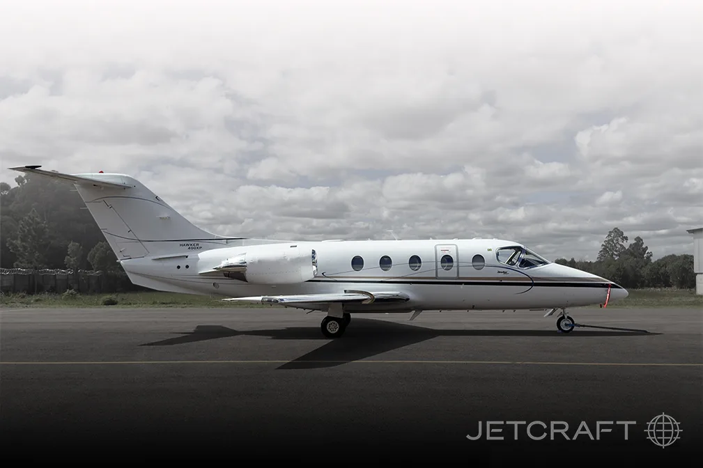 A white Hawker 1000XP business jet with dark pinstripes on the fuselage is parked on an asphalt runway under a cloudy sky.