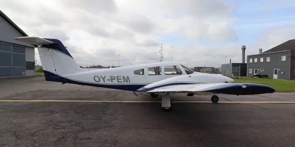 A white single-engine propeller aircraft with blue trim, registration OY-PEM, parked on an asphalt tarmac next to a hangar under a cloudy sky.