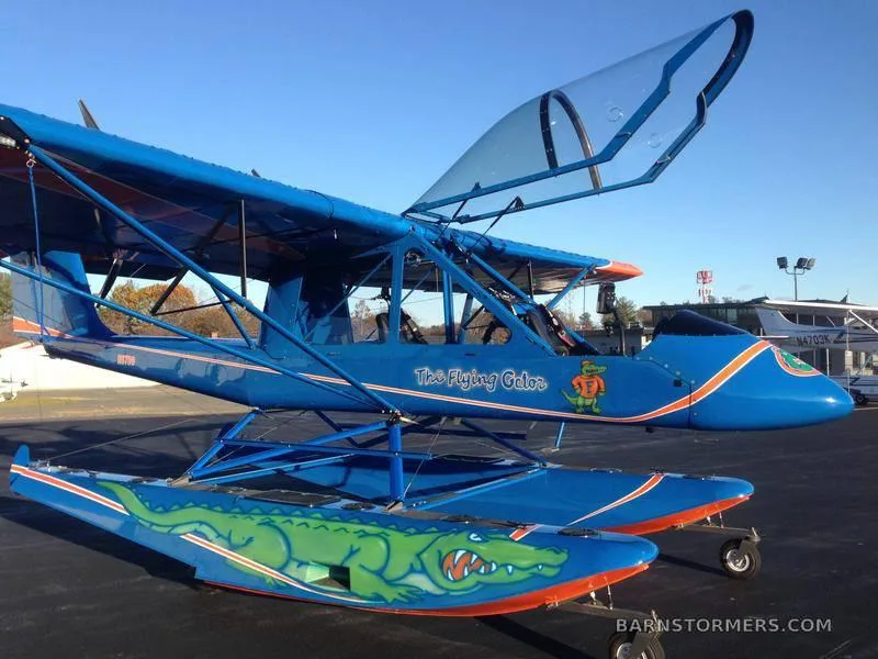 A blue floatplane with "The Flying Gator" painted on its side and a large green alligator graphic on its floats, parked on an tarmac.