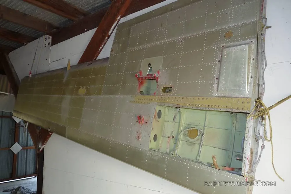 A close-up view of a riveted metal aircraft wing or tail section, showing internal structure, likely in a hangar.