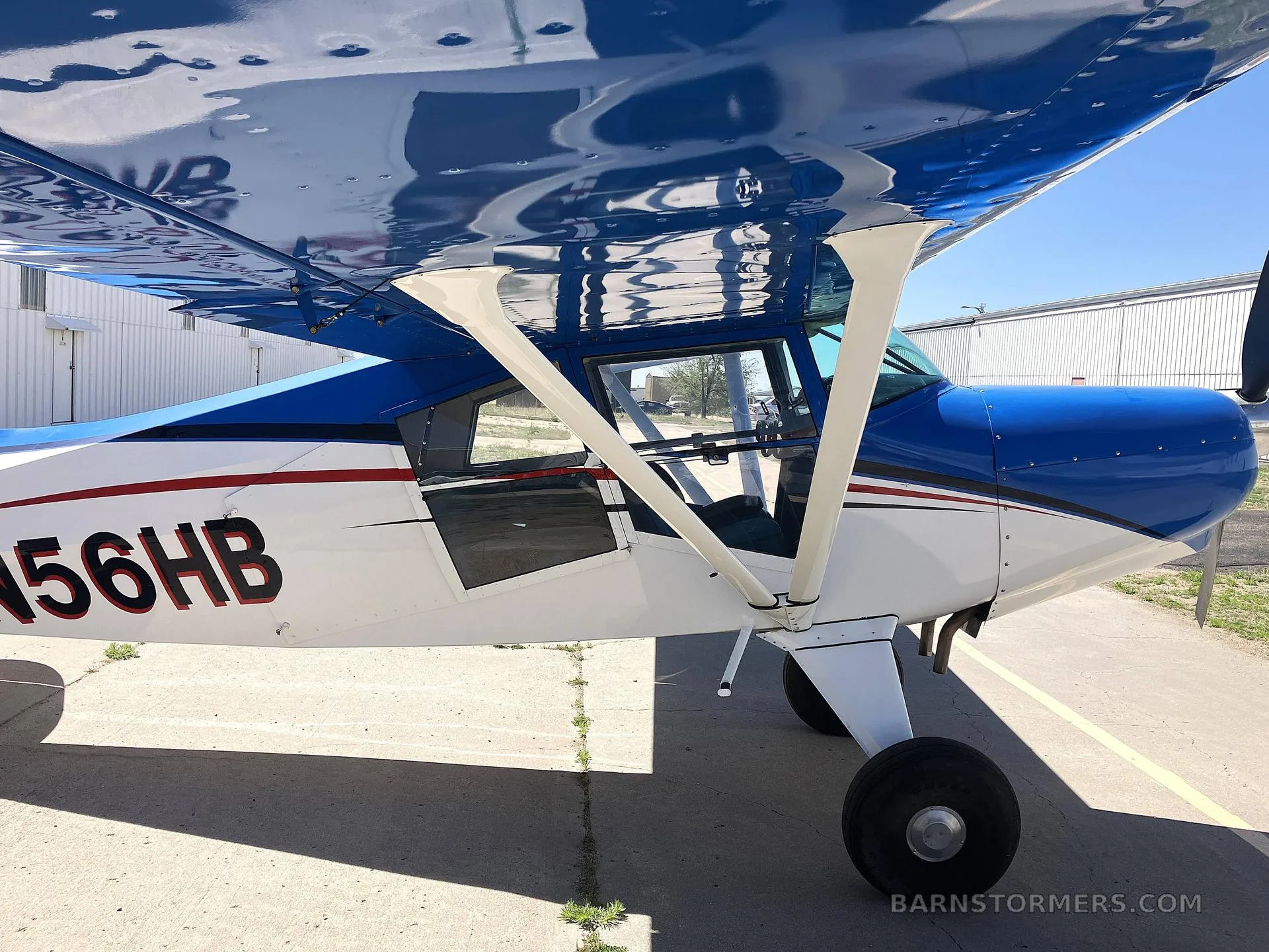 A close-up side view of the cabin and wing of a blue and white high-wing aircraft, registration N56HB, parked on an apron.