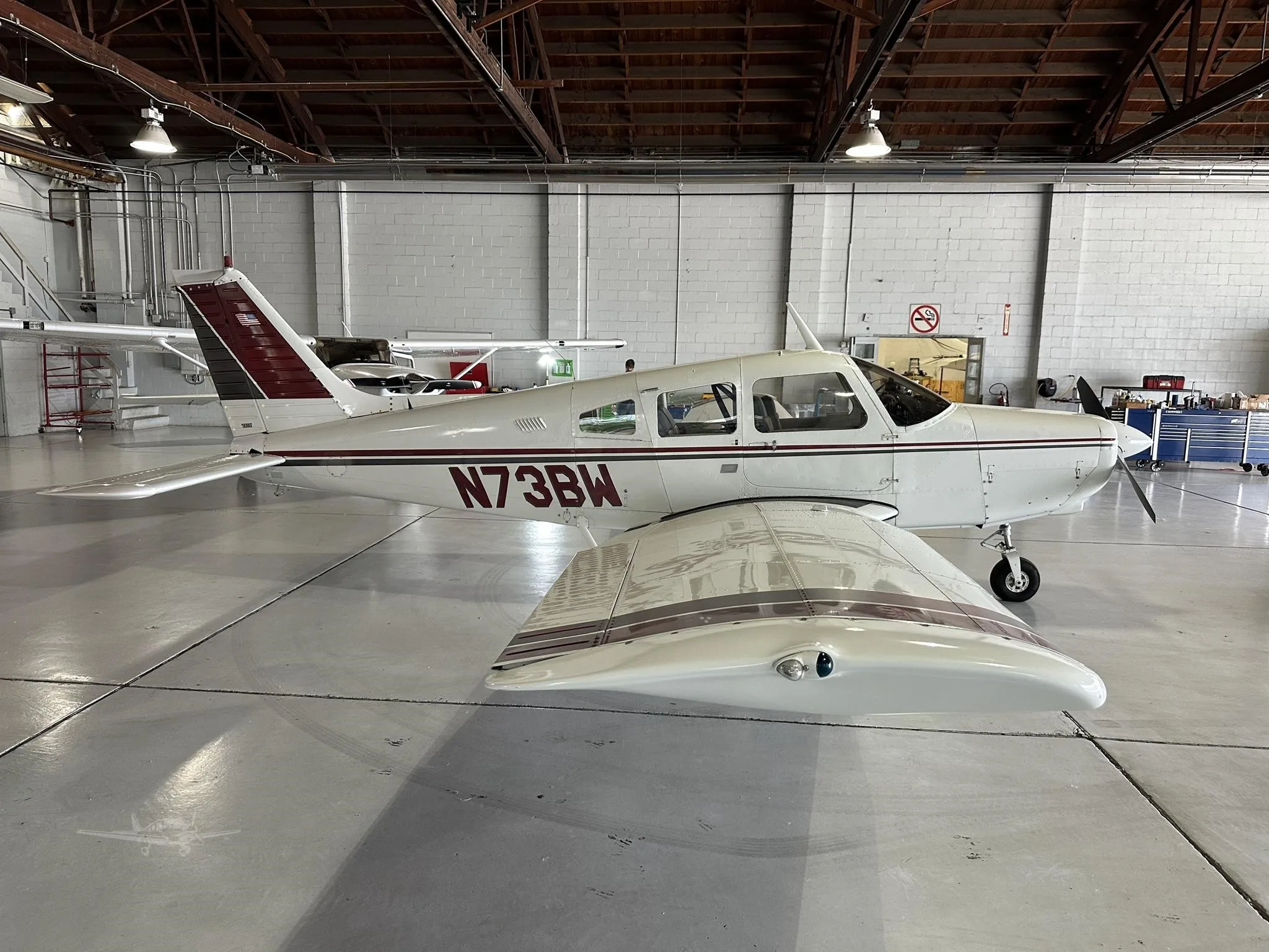 A white single-engine aircraft with maroon and black stripes, registration N73BW, parked in a hangar.