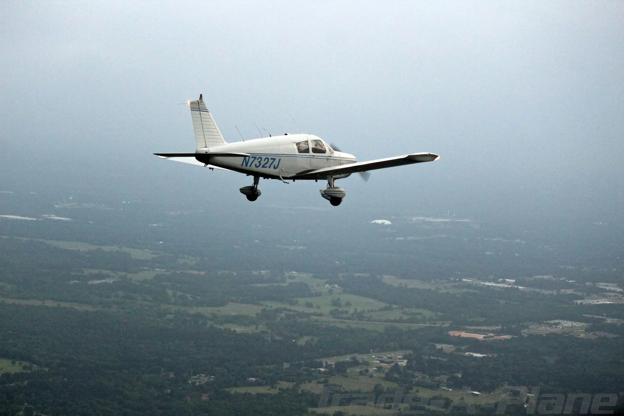 A white Piper Cherokee 140 aircraft, N7327J, with blue stripes, flies high above a green landscape under an overcast sky.