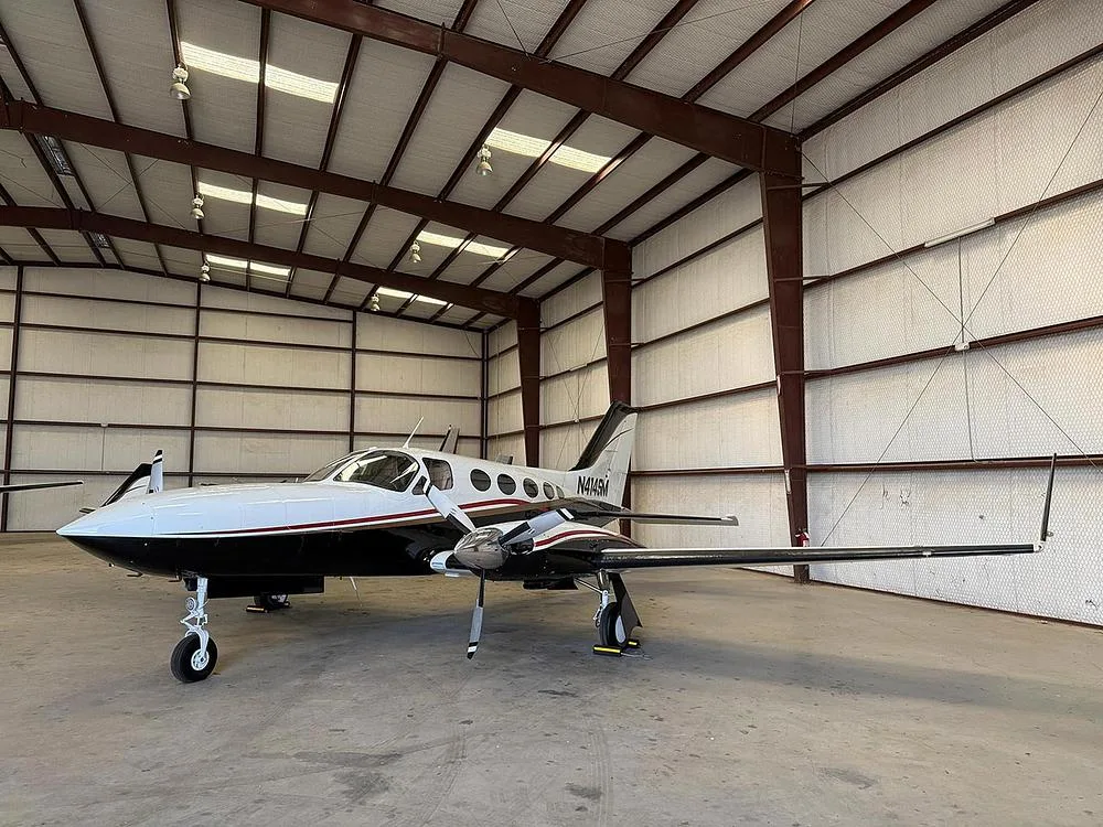 A white and black twin-engine aircraft, N414SM, with gold trim, parked inside a hangar, viewed from the front-left.