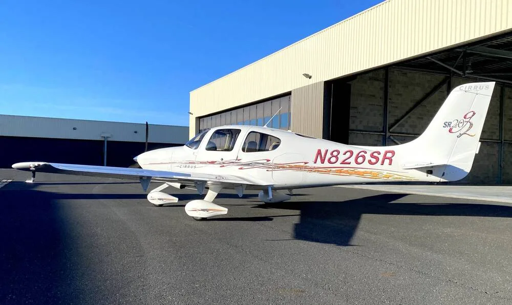 A side view of a white Cirrus SR22 aircraft with red and gold accent stripes, registration N826SR, parked on an airfield.