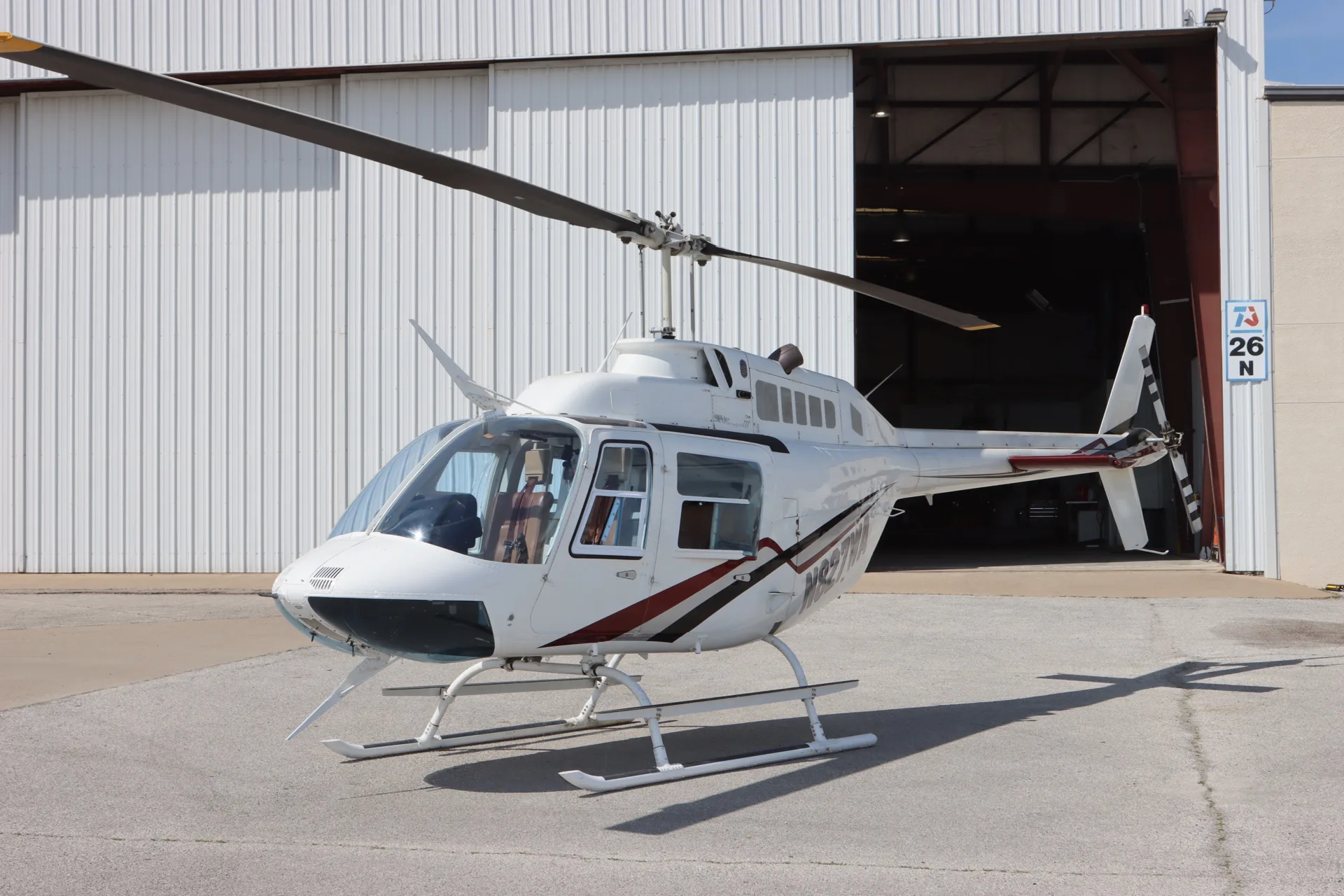 A white Bell 206 JetRanger helicopter with red and black stripes is parked on asphalt in front of an open hangar door.