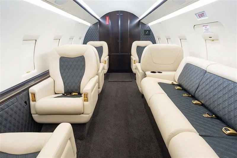 The spacious interior cabin of a business jet, featuring cream and dark grey quilted seats and a long sofa.