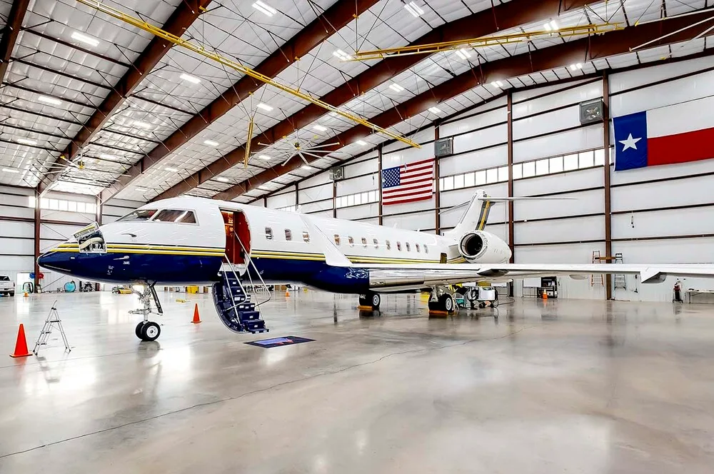 A Bombardier Challenger 605 business jet with blue and white livery parked inside a hangar with an open door and stairs.