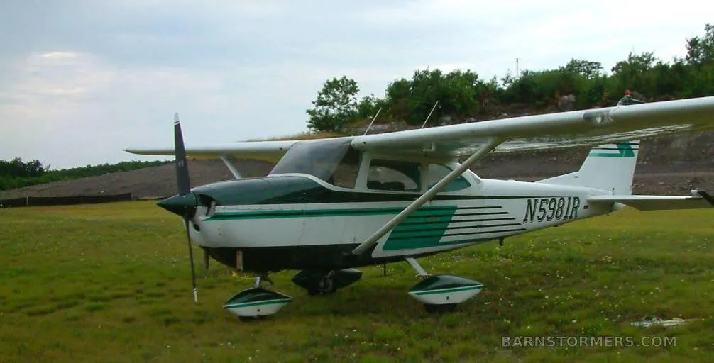 A white and green Cessna N5981R parked on a grassy field under a cloudy sky.