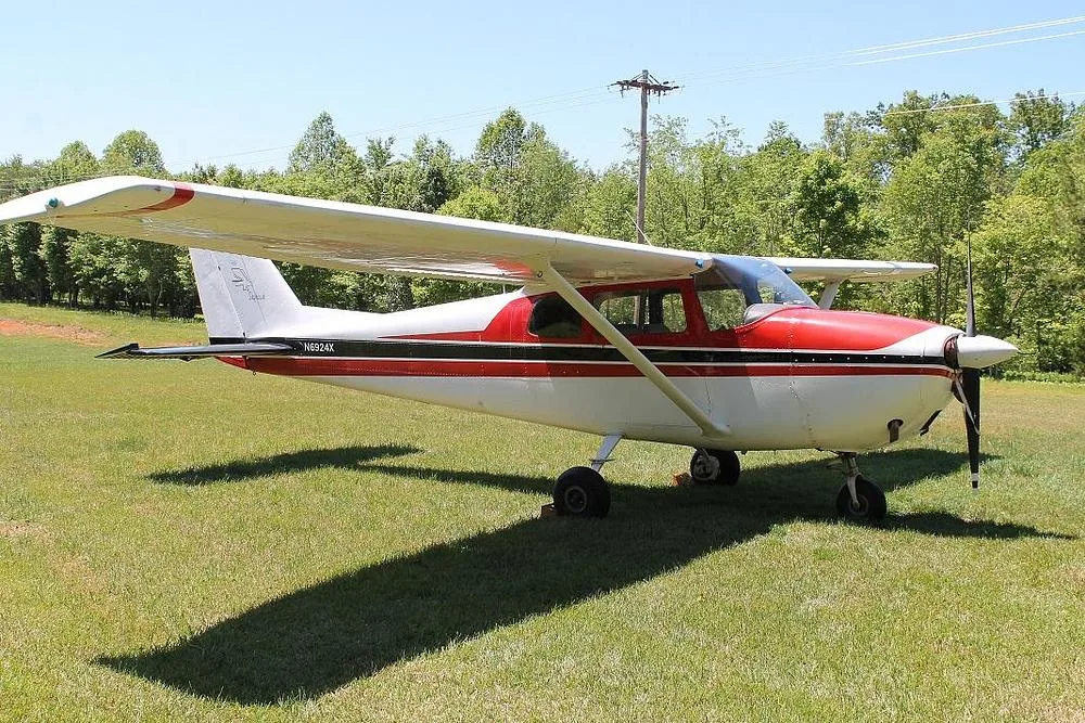 A side view of a red and white Cessna 172 (N6924X) with a black stripe, casting a long shadow on a grassy field.