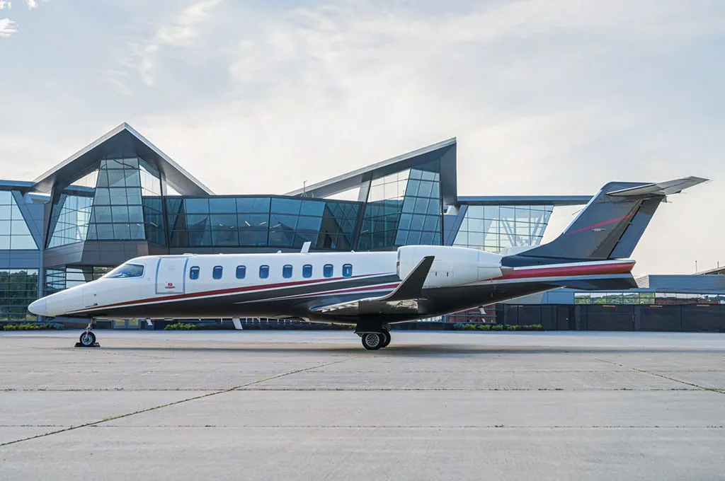 A Learjet 75 business jet with a white, black, and red paint scheme parked on an airport tarmac in front of a modern glass building.