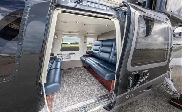 The open side door of a dark grey helicopter reveals its luxurious interior cabin with blue leather seating and a patterned carpet.