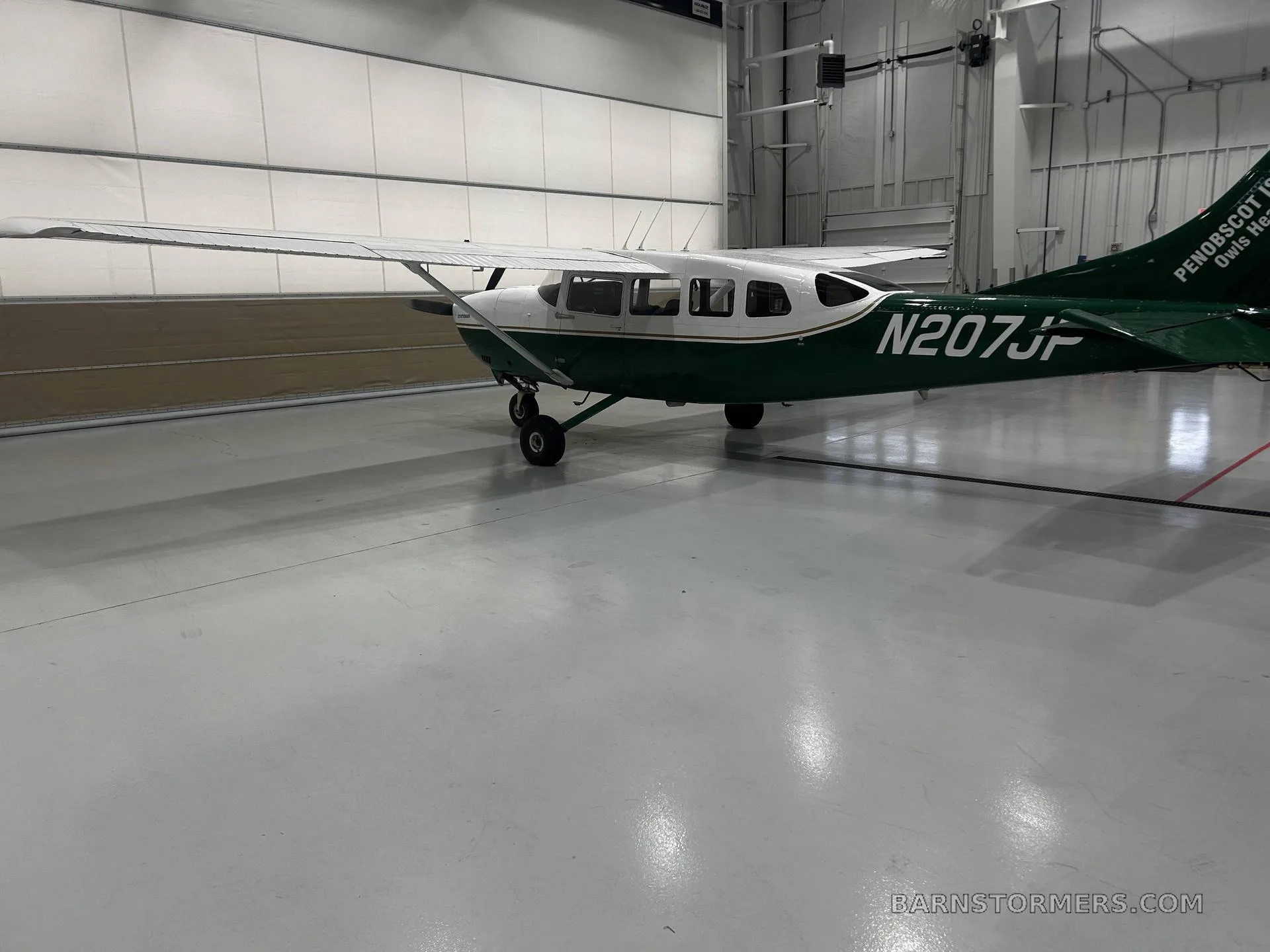 A white and green Cessna 207 Stationair, registration N207JP, is parked in a hangar, viewed from the rear-right side.