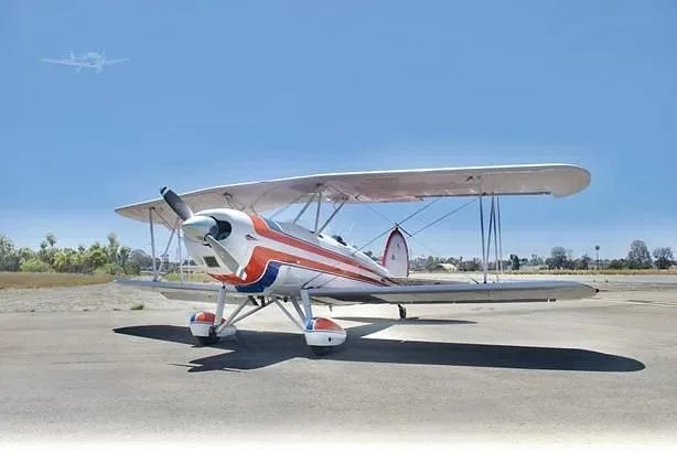 A white biplane with red and blue stripes parked on an airfield under a clear blue sky.