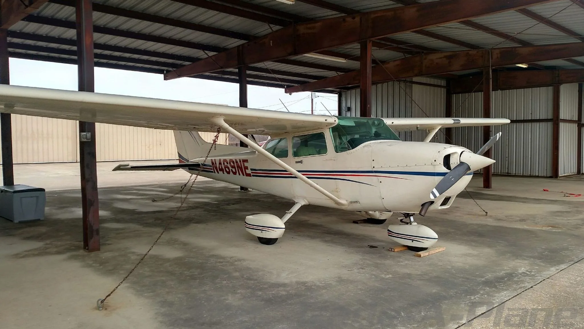 A white Cessna 172 with blue and red stripes parked under a metal hangar.