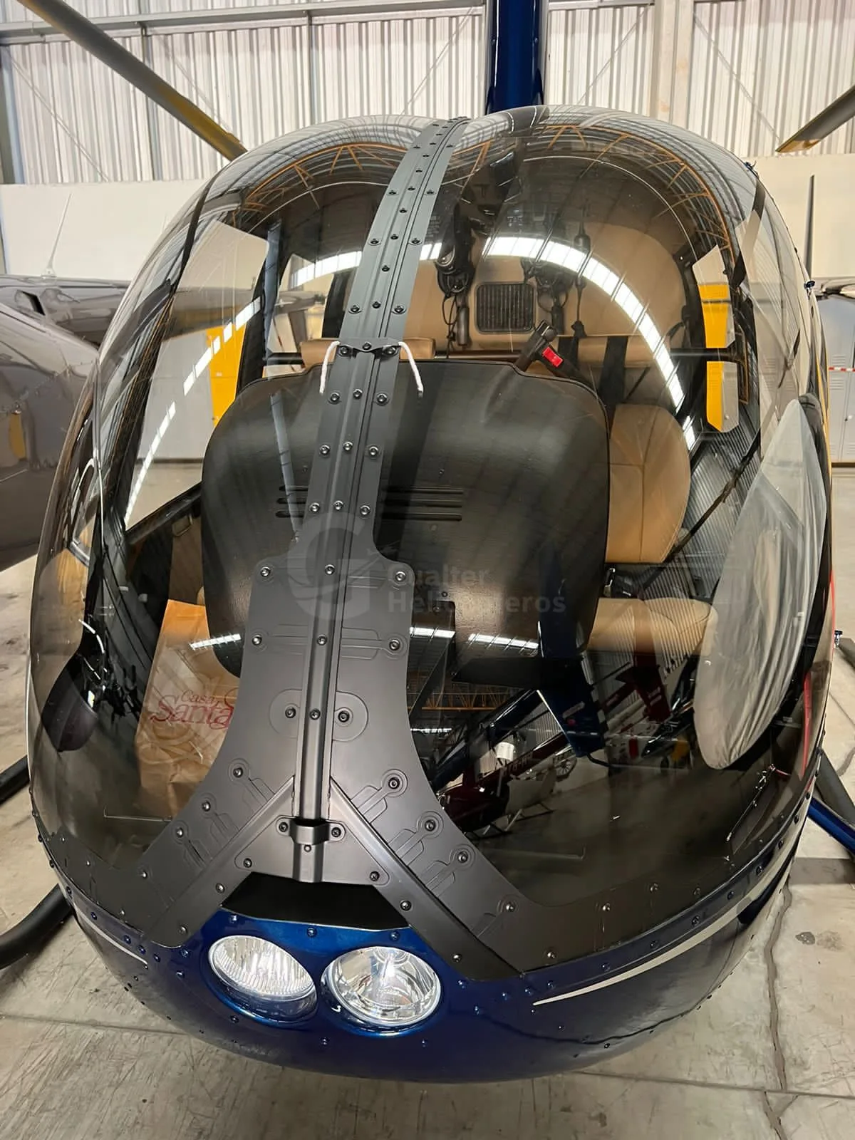 A close-up front view of a blue Robinson R44 Raven II helicopter, showing its clear bubble windshield and tan interior seats.