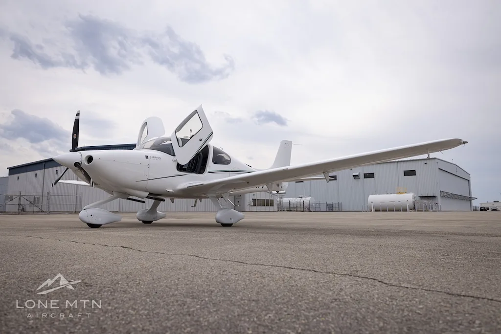 A white Cirrus SR22 aircraft with green accent stripes and open doors, parked on an asphalt ramp.