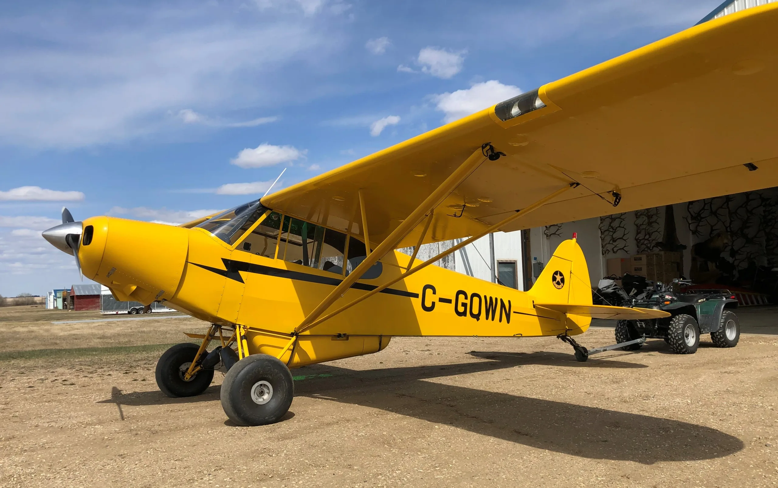 A bright yellow Piper Cub-style aircraft with large tundra tires, registration C-GQWN, is parked on a dirt field next to a hangar with an ATV.