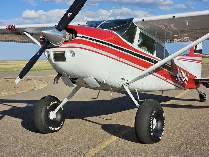 A red and white Cessna 185 Skywagon with large tundra tires, parked on an airfield under a blue sky.